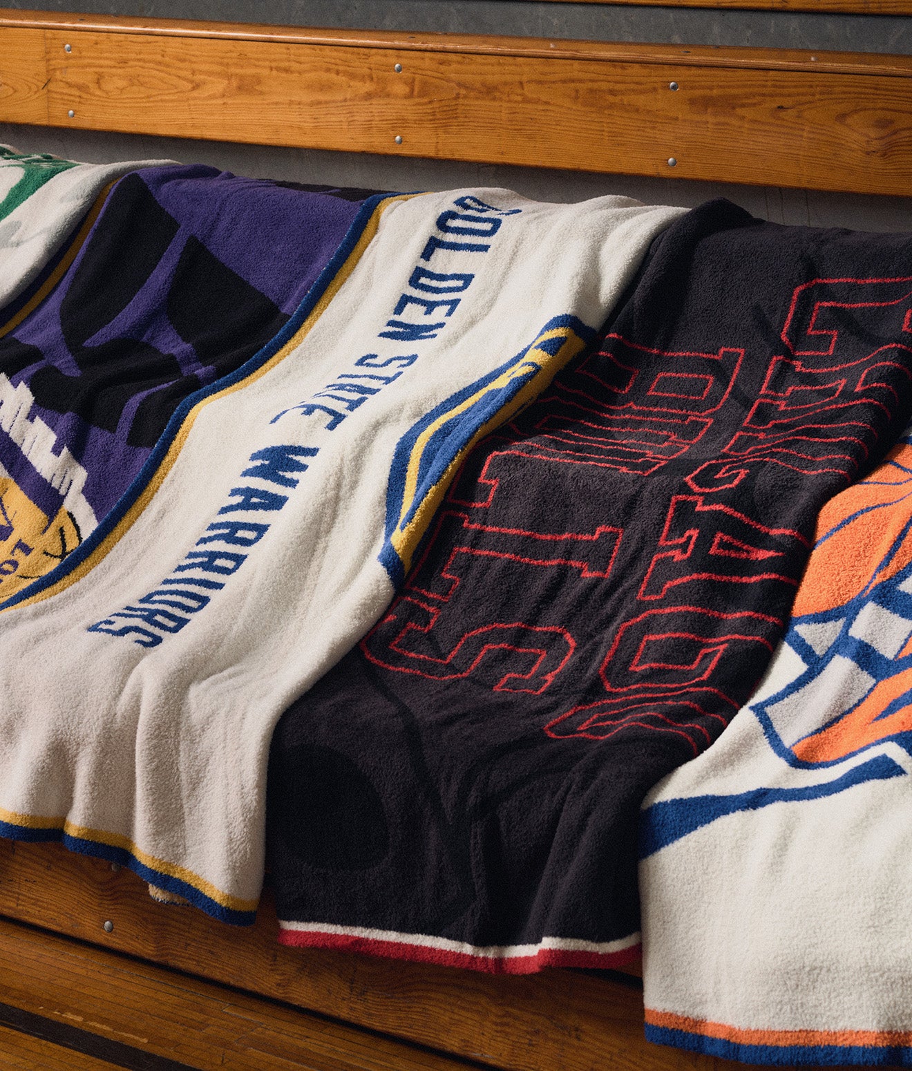 Four woven sports blankets draped over a wooden bench, featuring team designs and text including 'Golden State Warriors' and 'Chicago Bulls'.