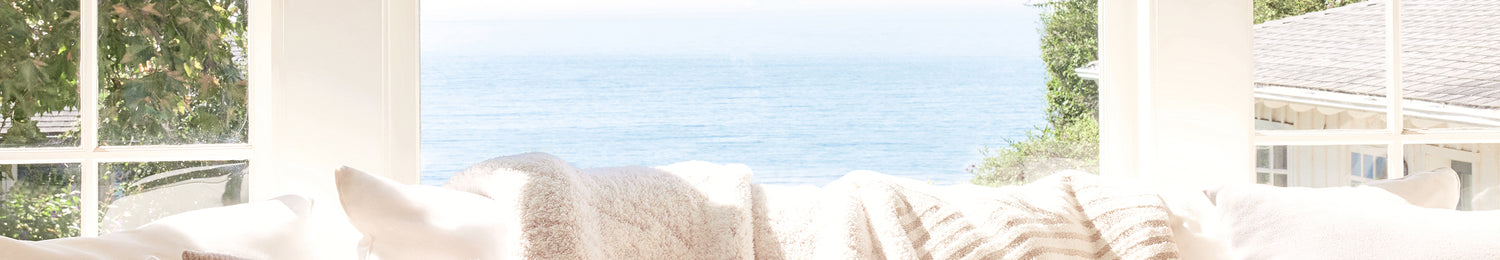 Cozy living room with white cushions on a sofa, large windows, and a view of greenery outside.
