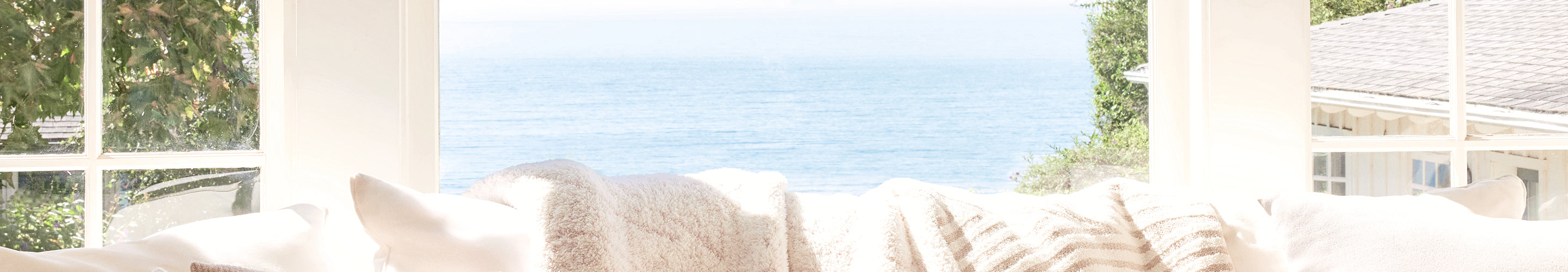 Cozy living room with white cushions on a sofa, large windows, and a view of greenery outside.