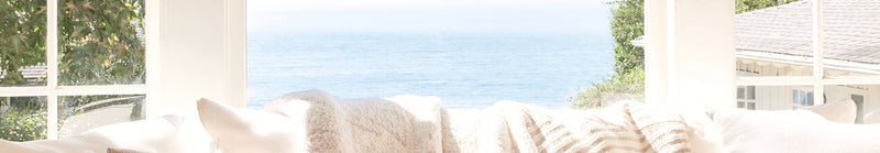 Cozy living room with white cushions on a sofa, large windows, and a view of greenery outside.