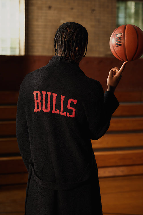 Caleb Wilson seen from behind wearing a black hoodie with 'BULLS' in red lettering on the back, holding a basketball in an indoor gym setting with wooden bleachers and brick walls.