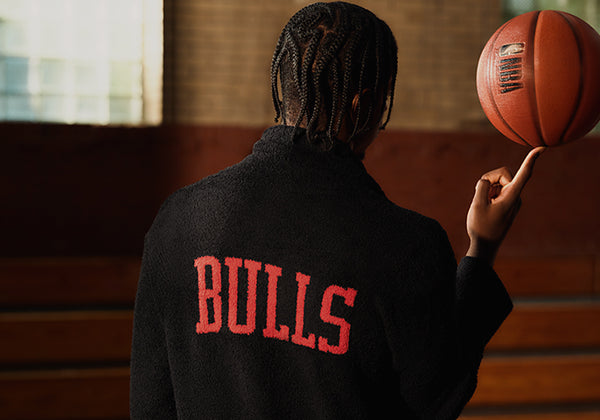 Caleb Wilson seen from behind wearing a black textured sweatshirt with 'BULLS' in red lettering on the back, balancing a basketball on their finger in an indoor gymnasium.